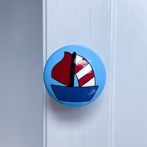 May include: A blue ceramic knob featuring a painted sailboat design. The sailboat has a red sail with white stripes and a blue hull.