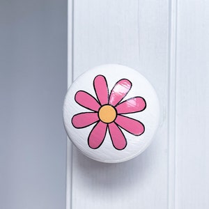 May include: White round cabinet knob with a pink flower design. The flower has a yellow centre and black outlines.