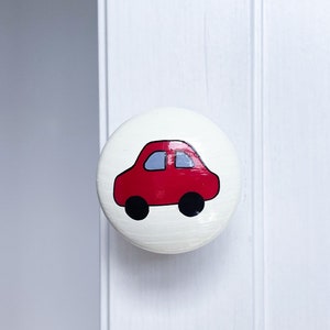 May include: A white round knob with a red car painted on it. The car has black wheels and a grey windscreen.