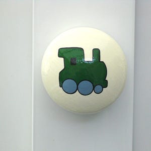 May include: A white round knob with a green train painted on it. The train has blue wheels and a black window.