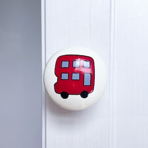 May include: A white ceramic knob featuring a red double-decker bus design. The bus has grey windows and black wheels.