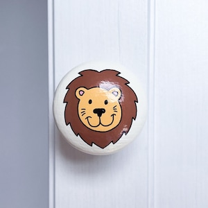May include: A white round knob with a painted lion face. The lion has brown fur, a yellow face, and pink ears.