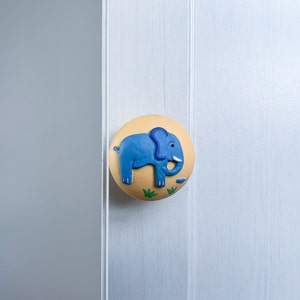 May include: A yellow and blue ceramic door knob featuring a cartoon elephant design. The elephant is blue with a light blue trunk and ears.