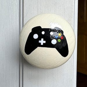 May include: A white round knob with a black video game controller painted on it. The controller has white buttons and a white plus sign. The buttons are coloured red, green, blue, and yellow.