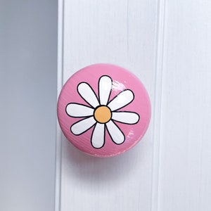 May include: Pink round wooden knob with a white daisy painted on it. The daisy has a yellow centre.