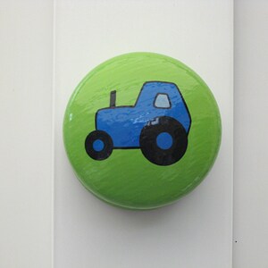 May include: A circular, green cabinet knob featuring a blue tractor illustration. The tractor has black wheels and a blue body. The knob is set against a white background.