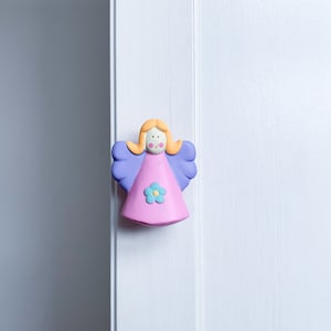 May include: A pink and purple ceramic angel figurine with a yellow halo and a blue flower on its dress. The angel is attached to a white door.