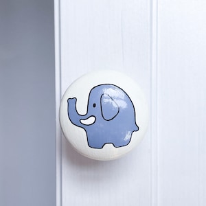 May include: A white ceramic knob featuring a blue elephant design. The elephant has a white tusk and a white eye.
