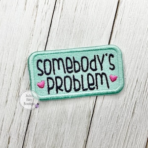 May include: A rectangular embroidered patch with a light mint green background and a dark green stitched border. The text reads SOMEBODY'S PROBLEM in black capital letters, accented by two small pink embroidered hearts on either side.