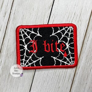 May include: A rectangular embroidered patch with a black background, a thick red border, and white spiderweb stitching. The text "I bite" is embroidered in red gothic-style lettering in the center, with a small red drop below the letter e.