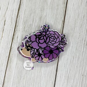 May include: A purple and black floral hedgehog-shaped acrylic pin. The pin features a light brown face and is adorned with various purple flowers. The pin has a glittery border and a small, round logo at the bottom.