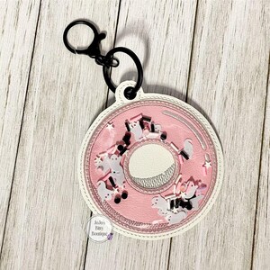 May include: Pink donut-shaped keychain with a white border and black hardware. The center of the donut is white, and the pink section contains small, decorative elements. The keychain is attached to a black clasp.
