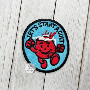 May include: Embroidered patch featuring a red cartoon character with white hair and horns, running with arms outstretched. The patch has a black border and a light blue background with the text "LET'S START A CULT".