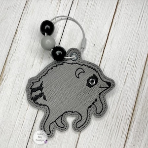 May include: A gray embroidered raccoon keychain featuring black stitched details, a striped tail, and a circular metal ring adorned with two black beads and one gray bead. The keychain includes a small circular tag labeled JoJo's Tiny Boutique.