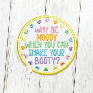 May include: A circular embroidered patch with a white background and a yellow border. The text reads WHY BE MOODY WHEN YOU CAN SHAKE YOUR BOOTY? in multi-colored letters, surrounded by small embroidered hearts in pink, blue, green, and yellow.