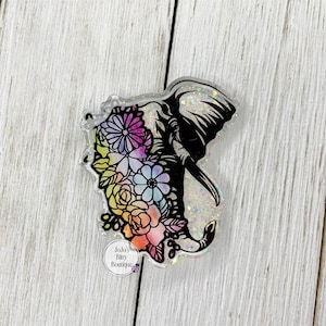 May include: A clear, glittery acrylic elephant-shaped charm. The elephant is black with a floral design in shades of pink, purple, yellow, and blue. The charm has a small, circular logo that reads "Jojo's Bay Boutique."