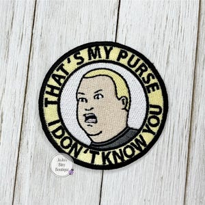 May include: Embroidered patch featuring a cartoon character with a surprised expression. The patch is circular, with the text "THAT'S MY PURSE! I DON'T KNOW YOU!" in black lettering on a yellow background. The character's face is in the center.