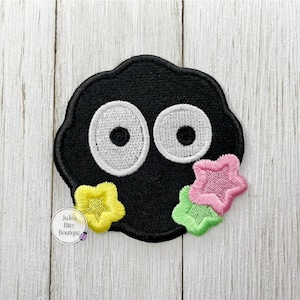 May include: A black embroidered patch featuring a cartoon-like character with large white eyes. The character has a rounded shape with yellow, pink, and green star accents. The patch is on a white wooden surface.