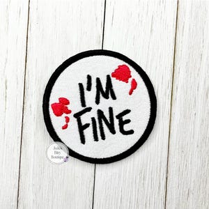 May include: Embroidered patch with the text "I'M FINE" in black, surrounded by a black border. Red blood-like splatters are on the white background. A fun, humorous accessory.