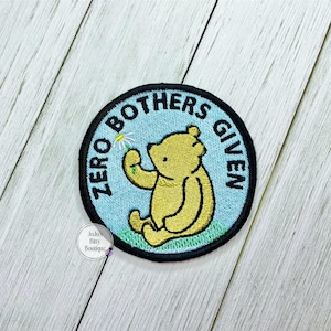 May include: Embroidered patch featuring a yellow bear holding a daisy, set against a light blue background. The patch is round with the words "ZERO BOTHERS GIVEN" in black lettering. The bear is sitting on green grass.