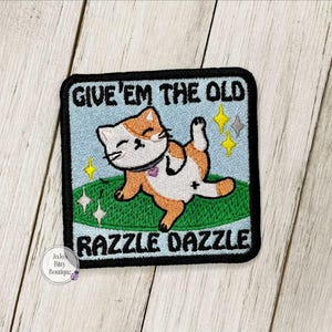May include: A square embroidered patch with a black border featuring a light blue background, a green oval patch of grass, and an orange and white cat posing. The text reads GIVE 'EM THE OLD RAZZLE DAZZLE, accompanied by yellow and white star icons.