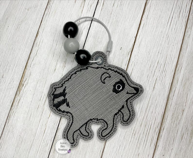 May include: A gray embroidered raccoon keychain featuring black stitched details, a striped tail, and a circular metal ring adorned with two black beads and one gray bead. The keychain includes a small circular tag labeled JoJo's Tiny Boutique.