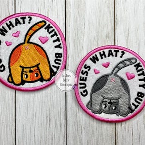 May include: Two round embroidered patches with a cat design. One patch features an orange cat, the other a gray cat, both with their rear ends facing up. The patches have the text "Guess What? Kitty Butt" in black and pink.