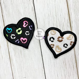 May include: Two heart-shaped patches. One is black with colorful heart and leopard print designs. The other is white with brown leopard print designs. Both have a black border and are on a light wood background.