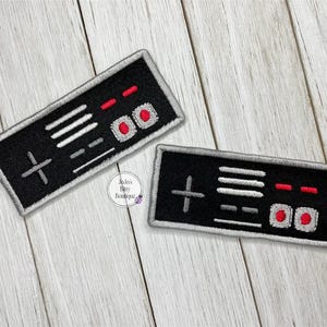 May include: Two black and white embroidered patches with a retro video game controller design. The patches have a gray border and red buttons.