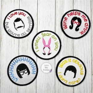 May include: Five embroidered patches with black borders and white centers. Each patch features a character from a cartoon show and a humorous phrase. The phrases include "I love you but you're all terrible" and "Wine helps me drink."