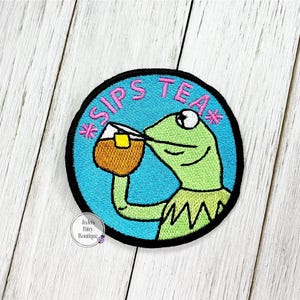 May include: Embroidered patch featuring a green frog drinking from a mug. The patch has a blue background with the words "SIPS TEA" in pink. The frog is wearing a green collar. The patch is round with a black border.