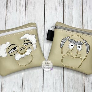 May include: Two beige zippered pouches with embroidered cartoon faces. The pouch on the left has a white face with a large nose and closed eyes. The pouch on the right has a tan face with a large nose and open eyes.