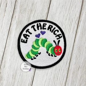 May include: Embroidered patch with a white background, black border, and the text "EAT THE RICH" in black. Features a green and yellow caterpillar with a red head and two purple hearts. The patch is round.