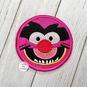 May include: Embroidered patch featuring a cartoon character's face with a pink circular border. The character has black fur, white eyes, a red nose, and a smiling mouth with white teeth. The patch includes the text "Jojo's Bitty Boutique".