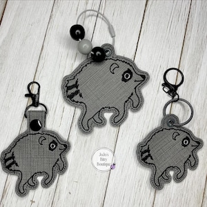 May include: Three gray embroidered raccoon keychains with black stitched details. One keychain features a silver wire loop with black and gray beads, while the other two have black metal lobster claw clasps and rings. A small circular logo reads Jojo's Baby Boutique.