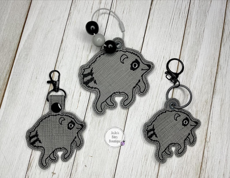 May include: Three gray embroidered raccoon keychains with black stitched details. One keychain features a silver wire loop with black and gray beads, while the other two have black metal lobster claw clasps and rings. A small circular logo reads Jojo's Baby Boutique.