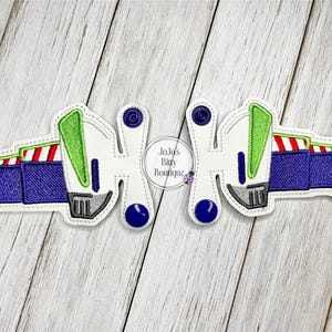 May include: Two embroidered patches in the shape of Buzz Lightyear's wings. The patches feature white, green, red, blue, and gray colors. The text "JoJo's Bitty Boutique" is in the center.