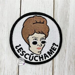 May include: Embroidered patch featuring a cartoon woman with brown hair and sunglasses. The patch has a white background with a black border and the text "¿ESCÚCHAME?" in black. The patch is round.