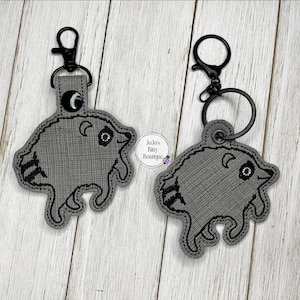 May include: Two gray embroidered keychains shaped like raccoons with black stitching details. Each keychain features a black metal lobster clasp and a circular ring attachment. The fabric has a textured weave pattern and includes a small logo tag.