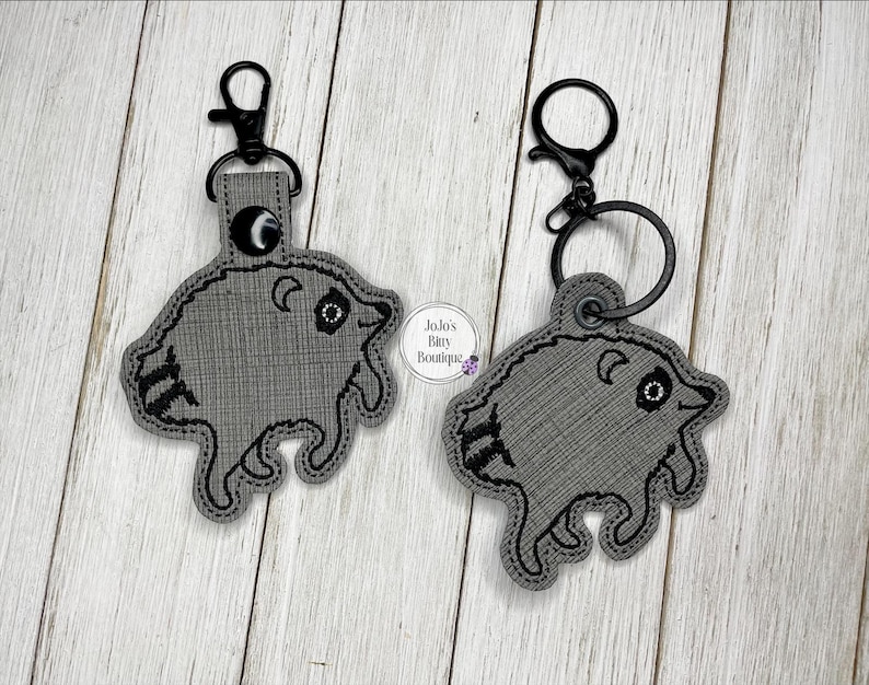 May include: Two gray embroidered keychains shaped like raccoons with black stitching details. Each keychain features a black metal lobster clasp and a circular ring attachment. The fabric has a textured weave pattern and includes a small logo tag.