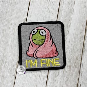 May include: Embroidered patch featuring a cartoon frog wrapped in a pink blanket. The frog has large, white-rimmed eyes and a green face. The text "I'M FINE" is embroidered in yellow. The patch has a black border and a gray background.