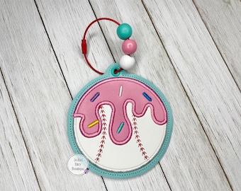 May include: A pink donut-shaped applique with white icing and colorful sprinkles, designed to resemble a baseball. It has a teal border and a red loop with teal, pink, and white beads. The product is from Jojo's Bitsy Boutique.