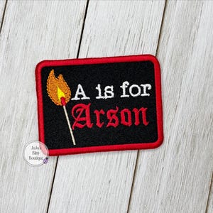 May include: Embroidered patch with a black background and a red border. The patch features a match with a flame and the words "A is for Arson" in white and red lettering.