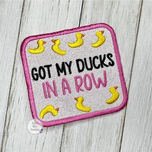 May include: Pink and white square patch with the text "GOT MY DUCKS IN A ROW" in black.  The patch is decorated with yellow rubber duck illustrations.