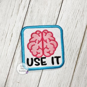 May include: A square embroidered patch with rounded corners featuring a white background, a bright blue border, a pink brain illustration in the center, and the black text USE IT positioned below the brain. The patch has a visible stitched edge.