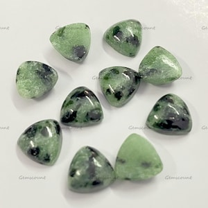 Peut inclure: Une collection de pierres précieuses triangulaires polies. Les pierres sont principalement vert clair avec des inclusions vert foncé et noires. Les pierres précieuses sont disposées sur une surface blanche. Le texte "Gemscount" est visible.