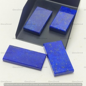May include: Four rectangular lapis lazuli gemstones with gold flecks are displayed. The stones are a deep blue color and are arranged on a white surface and a black tray. The gemstones are likely for jewelry making.