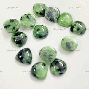May include: A selection of polished green and black zoisite gemstones. The stones are smooth and rounded, with a mix of light and dark green hues and black inclusions. The text "Gemscount" is printed on the white surface.