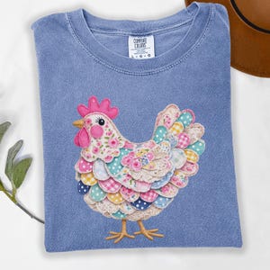 May include: A blue Comfort Colors t-shirt with a patchwork chicken design. The chicken has a pink comb and wattle, and its feathers are patterned with florals and polka dots. The shirt has a crew neck and short sleeves.