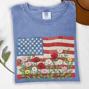 May include: A blue Comfort Colors t-shirt with an American flag design and embroidered flowers. The flag has a patchwork design with stars and stripes, and the flowers include poppies and daisies. The shirt is a size L-G-G.